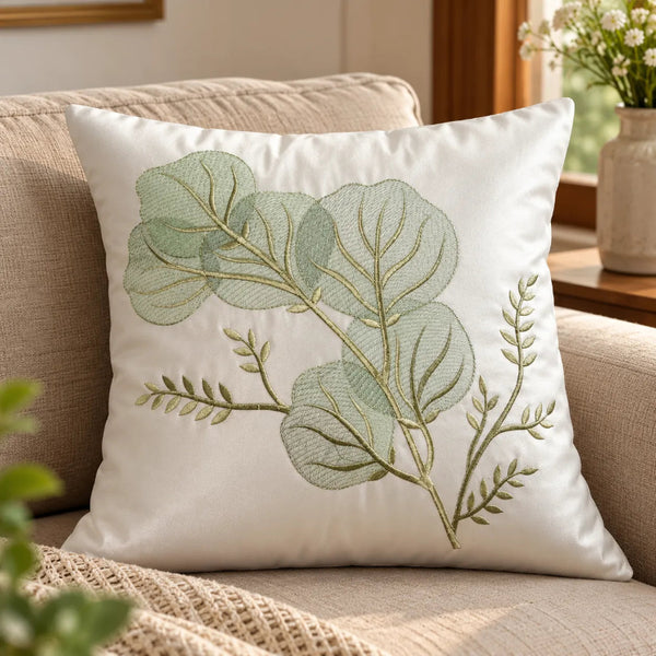 A square white throw pillow with an embroidered botanical design of green leaves and stems, resting on a beige upholstered sofa in a warm, sunlit room.