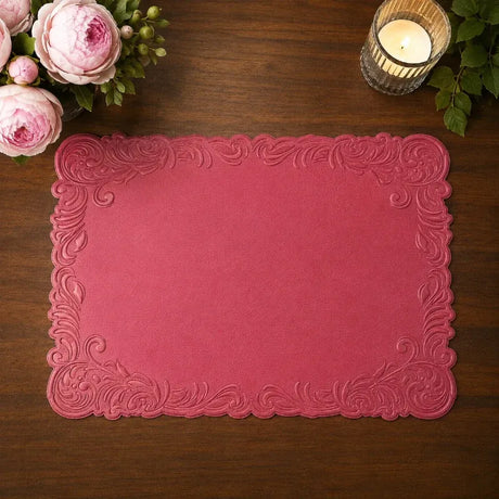 A rectangular pink placemat with an embossed ornate baroque-style border, displayed on a dark wooden table with pink roses and a lit candle in the corners.