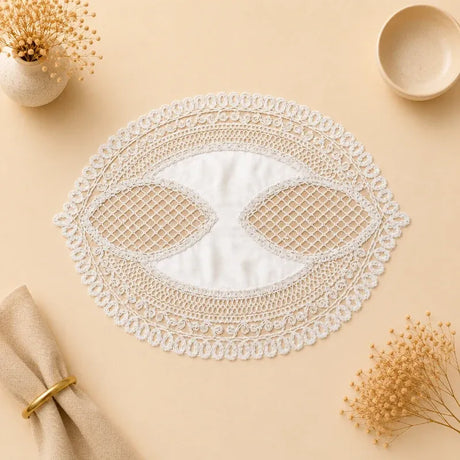 An oval white lace doily with intricate floral patterns and mesh cutouts, styled on a beige surface with dried flowers, a napkin, and a small bowl.