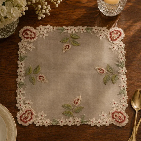 A square beige linen placemat with an embroidered floral border and scattered floral motifs, styled on a dark wooden table with a gold spoon, glass candle holder, and flowers.
