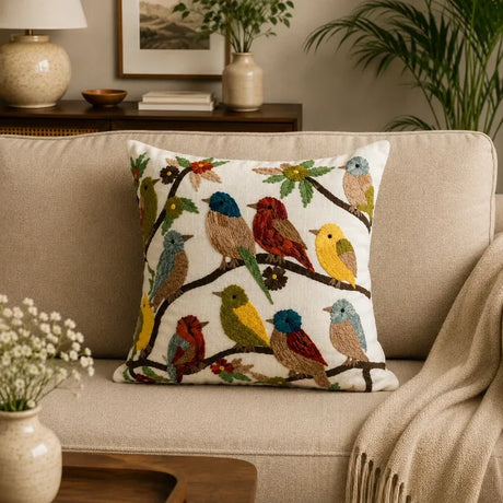 A square throw pillow with a detailed embroidered pattern of various colorful birds perched on branches, displayed on a beige fabric sofa in a cozy living room setting.