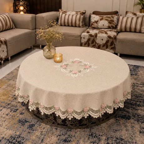 A round beige tablecloth with a white lace floral border and embroidered floral center, draped over a round coffee table in a living room setting.