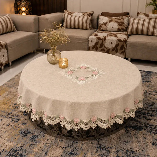 A round beige tablecloth with a white lace floral border and embroidered floral center, draped over a round coffee table in a living room setting.