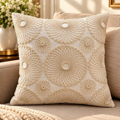 A square decorative throw pillow with intricate beige floral embroidery on a cream fabric background, styled on a beige sofa with a blurred background of flowers and a mirror.