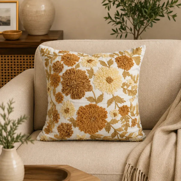 A square throw pillow with a textured floral pattern in mustard yellow, ochre, and cream on a white background, styled on a beige armchair with a throw blanket and home decor in the background.