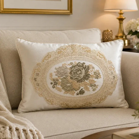 A square decorative throw pillow with an elegant embroidered floral design in beige and muted green on a cream satin background, styled on a beige sofa in a cozy living room setting.