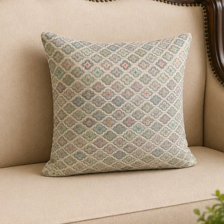 A square decorative throw pillow with a woven geometric quatrefoil pattern in muted blue, green, beige, and orange tones, resting on a beige fabric sofa.