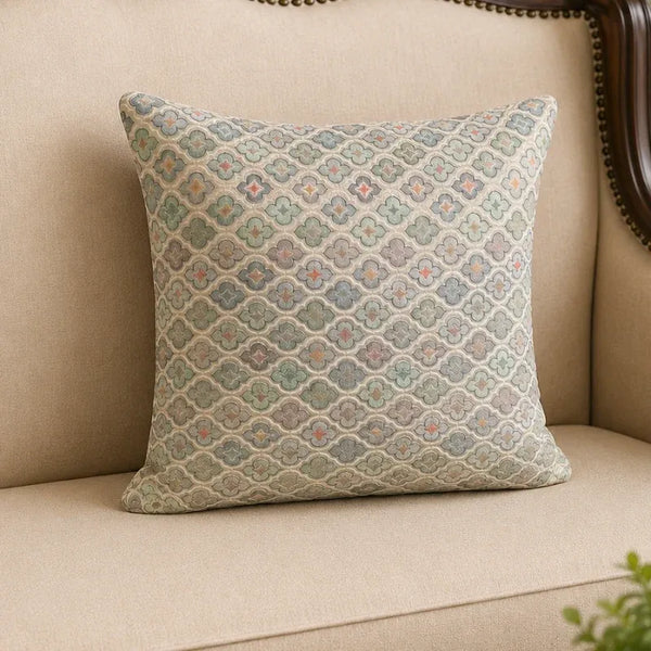A square decorative throw pillow with a woven geometric quatrefoil pattern in muted blue, green, beige, and orange tones, resting on a beige fabric sofa.