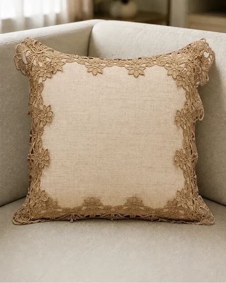 A square beige throw pillow with a natural linen-like center and a decorative floral lace border, resting on a light beige upholstered sofa.