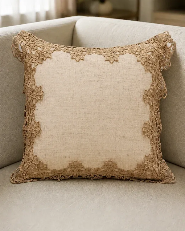 A square beige throw pillow with a natural linen-like center and a decorative floral lace border, resting on a light beige upholstered sofa.