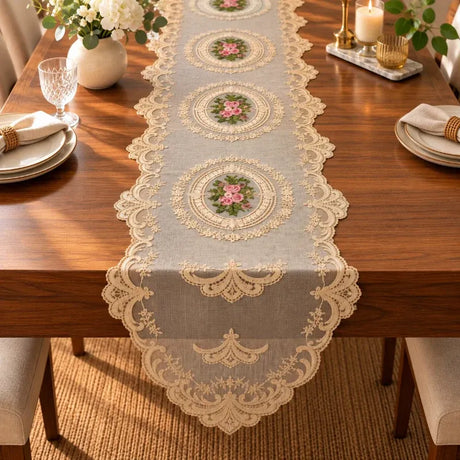 A light gray table runner with intricate cream lace trim and three central floral medallions, styled on a wooden dining table with plates, a vase of flowers, and candles.