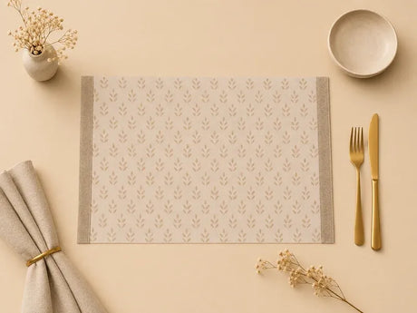 A rectangular beige placemat with a repeating leaf pattern and gray side borders, styled on a cream tabletop with a folded napkin, gold-toned fork and knife, a small bowl, and dried flowers.