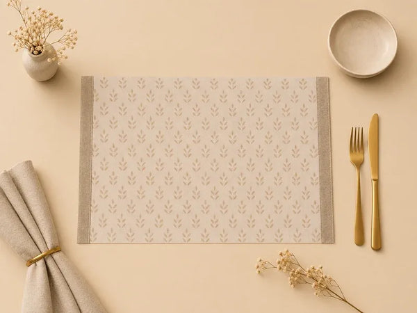 A rectangular beige placemat with a repeating leaf pattern and gray side borders, styled on a cream tabletop with a folded napkin, gold-toned fork and knife, a small bowl, and dried flowers.