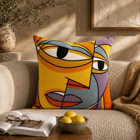 A square throw pillow with a colorful abstract face design in yellow, orange, blue, gray, and purple, styled on a beige textured sofa in a warm, sunlit living room.