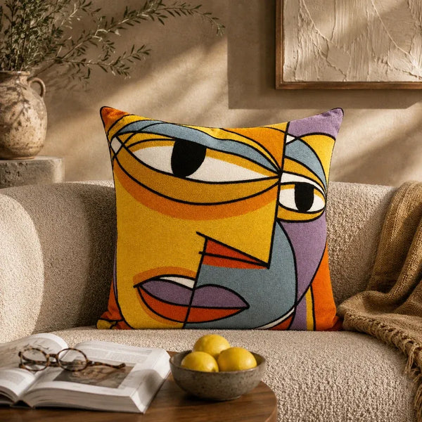 A square throw pillow with a colorful abstract face design in yellow, orange, blue, gray, and purple, styled on a beige textured sofa in a warm, sunlit living room.