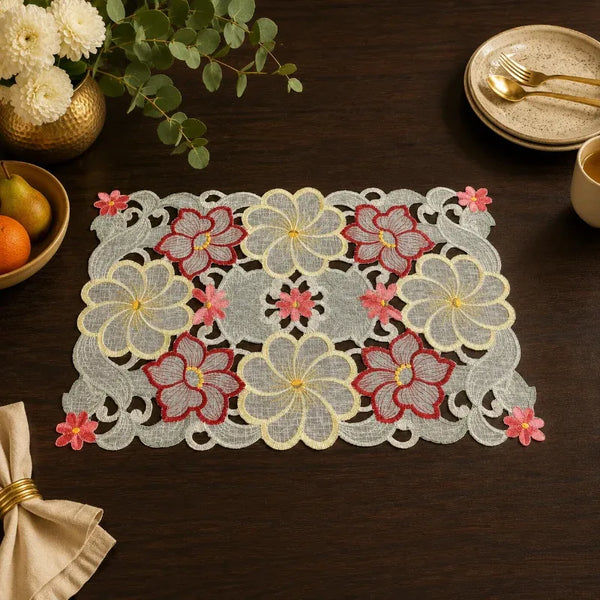 A rectangular embroidered lace placemat with floral motifs in pink, red, and cream on a white mesh base, styled on a dark wooden table with dining props.