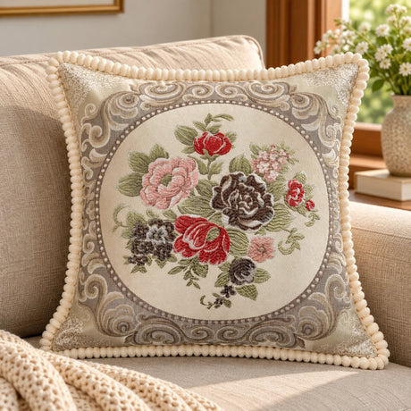Beige embroidered cushion cover with red, pink and brown floral design, decorative border and cream trim styled on a neutral sofa