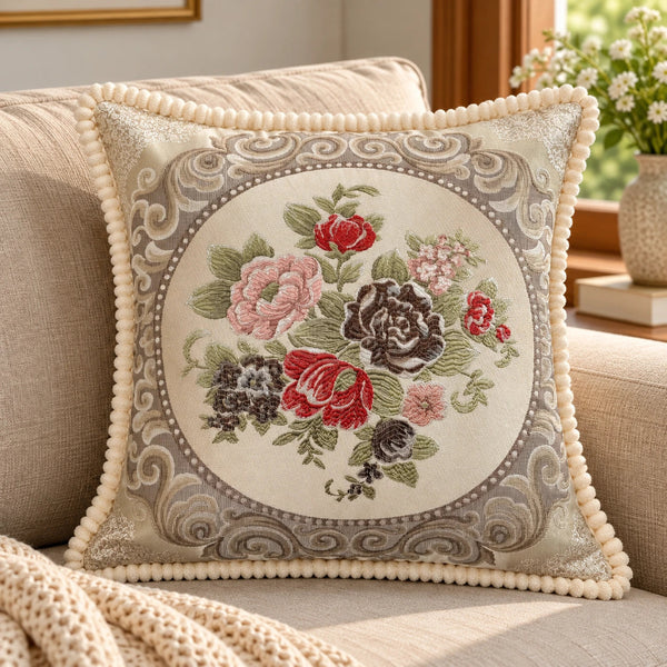 Beige embroidered cushion cover with red, pink and brown floral design, decorative border and cream trim styled on a neutral sofa